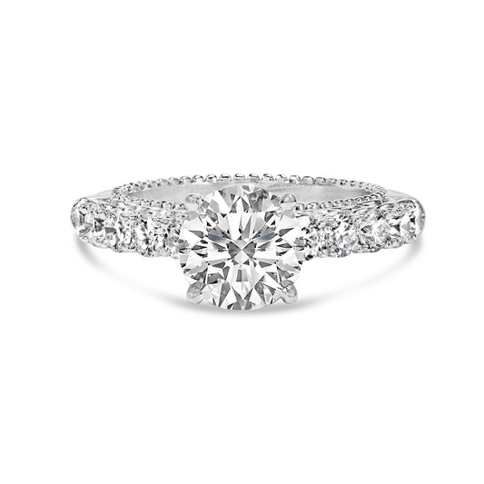 Front view of a white metal ring with one large round white gemstone in prong setting at the center and multiple smaller round white gemstones in shared prong setting along the band.