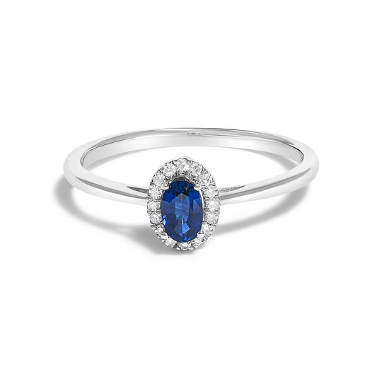 Close-up front angle of a white metal ring featuring a central oval blue gemstone surrounded by small round white gemstones in a micro-pave setting, shown on a white background.
